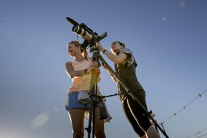 Two students setting up a video camera against a blue sky.